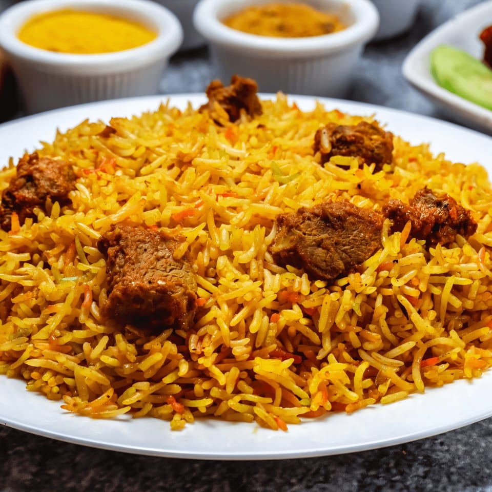 A white plate of golden spiced rice topped with grilled meat pieces, accompanied by small bowls of sauces and sliced cucumber.