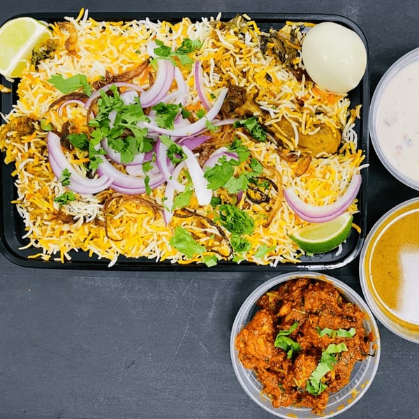 Chicken Biryani with lime, onion, cilantro, boiled egg, raita, and curry on a dark surface.